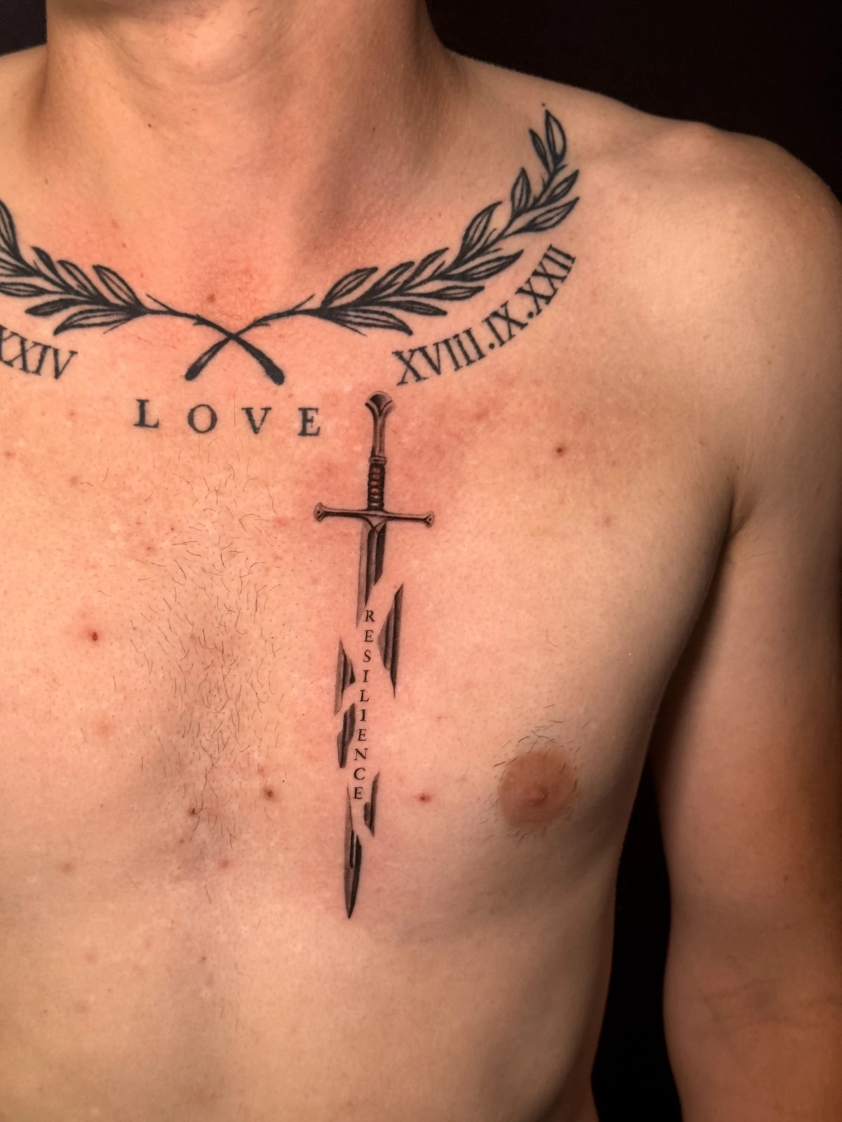 Tattoo mit Lorbeerzweigen, römischen Ziffern und schmalem Schwert mit dem Schriftzug Resilience auf der Brust – Arbeit von Merci, Skin City Tattoo.