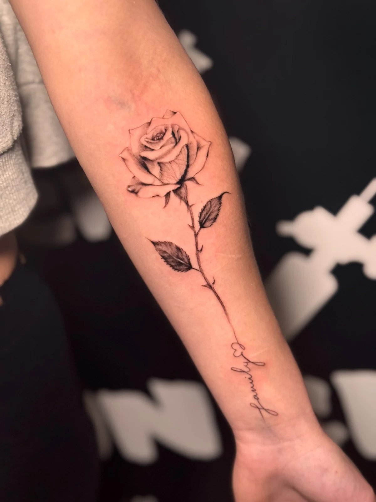 Fein schattierte Rose mit Stiel und geschwungenem Schriftzug am Unterarm – Arbeit von Merci, Skin City Tattoo.